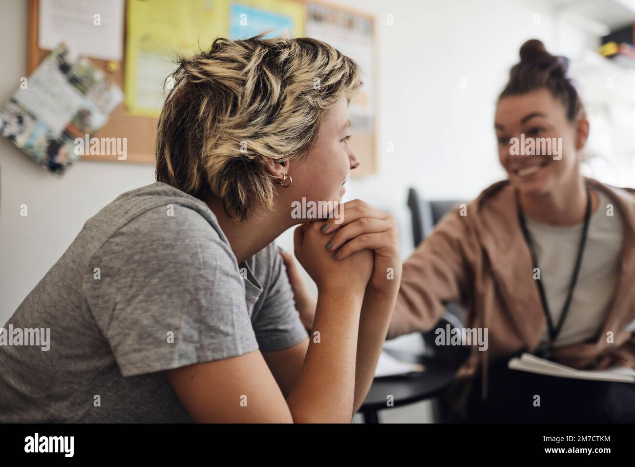 Professionnel de la santé mentale souriant regardant une étudiante assise avec la main sur le menton dans le bureau de l'école Banque D'Images