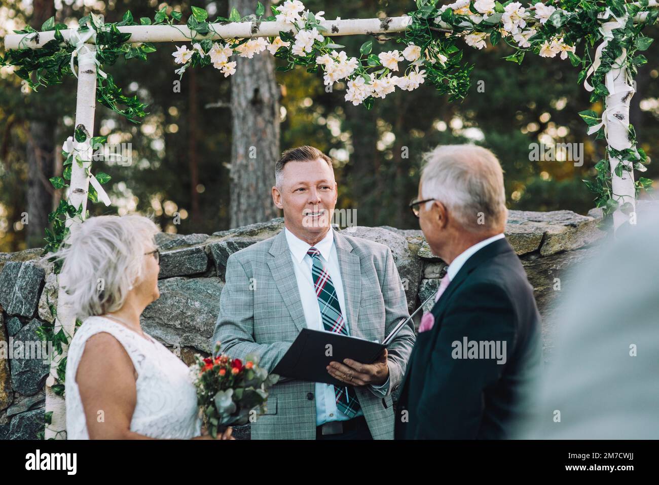 Couple senior échangeant des voeux tout en regardant le ministre debout contre le mur avec la décoration de fleur pendant le mariage céré Banque D'Images