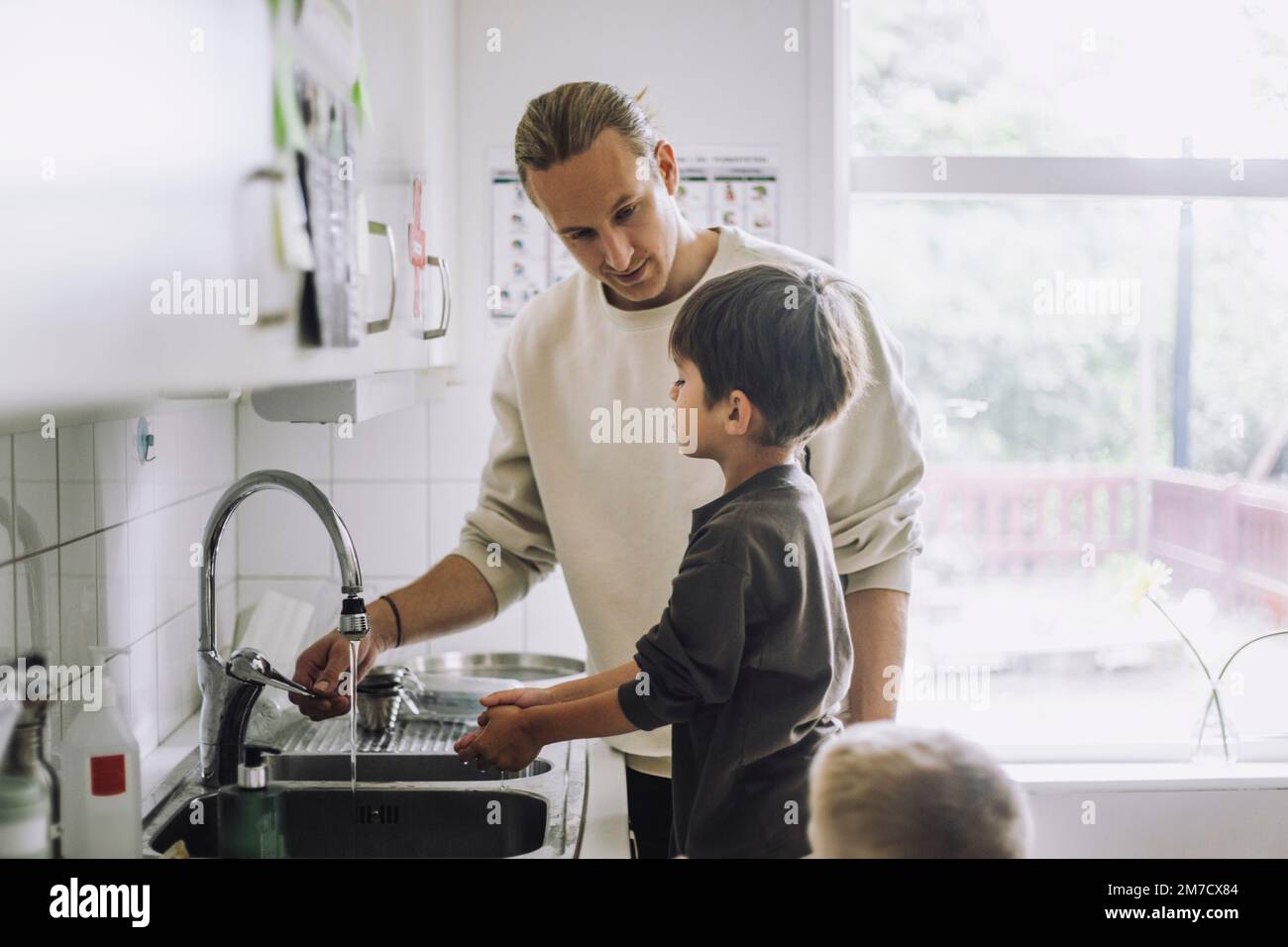 Un travailleur de garde d'enfants de sexe masculin enseigne au garçon de se laver les mains près de l'évier de la maternelle Banque D'Images