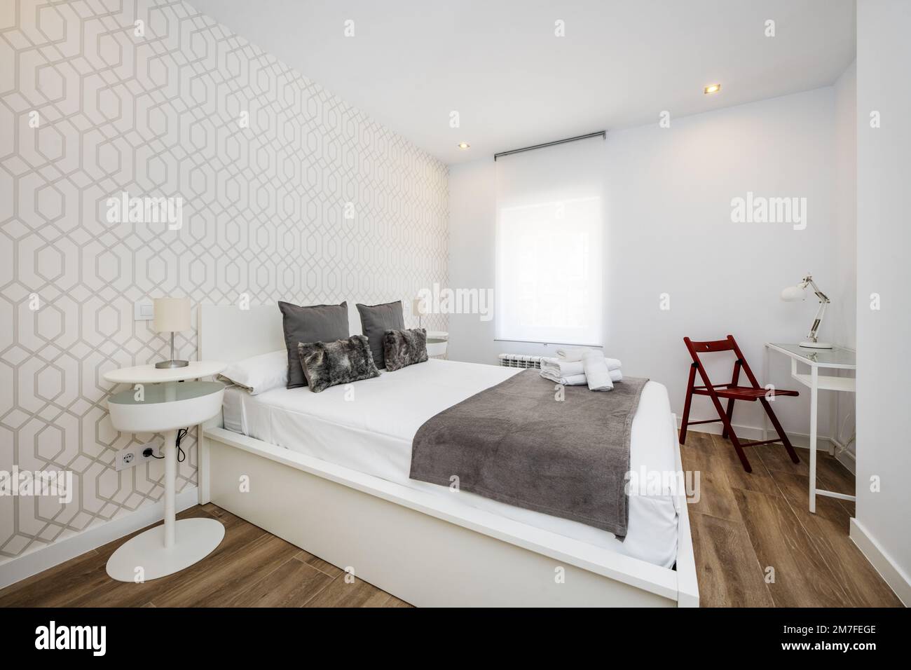 Chambre avec une tête de lit en bois blanc, des coussins gris, un lit en bois blanc et dans un coin un bureau avec une chaise pliante en bois rouge Banque D'Images