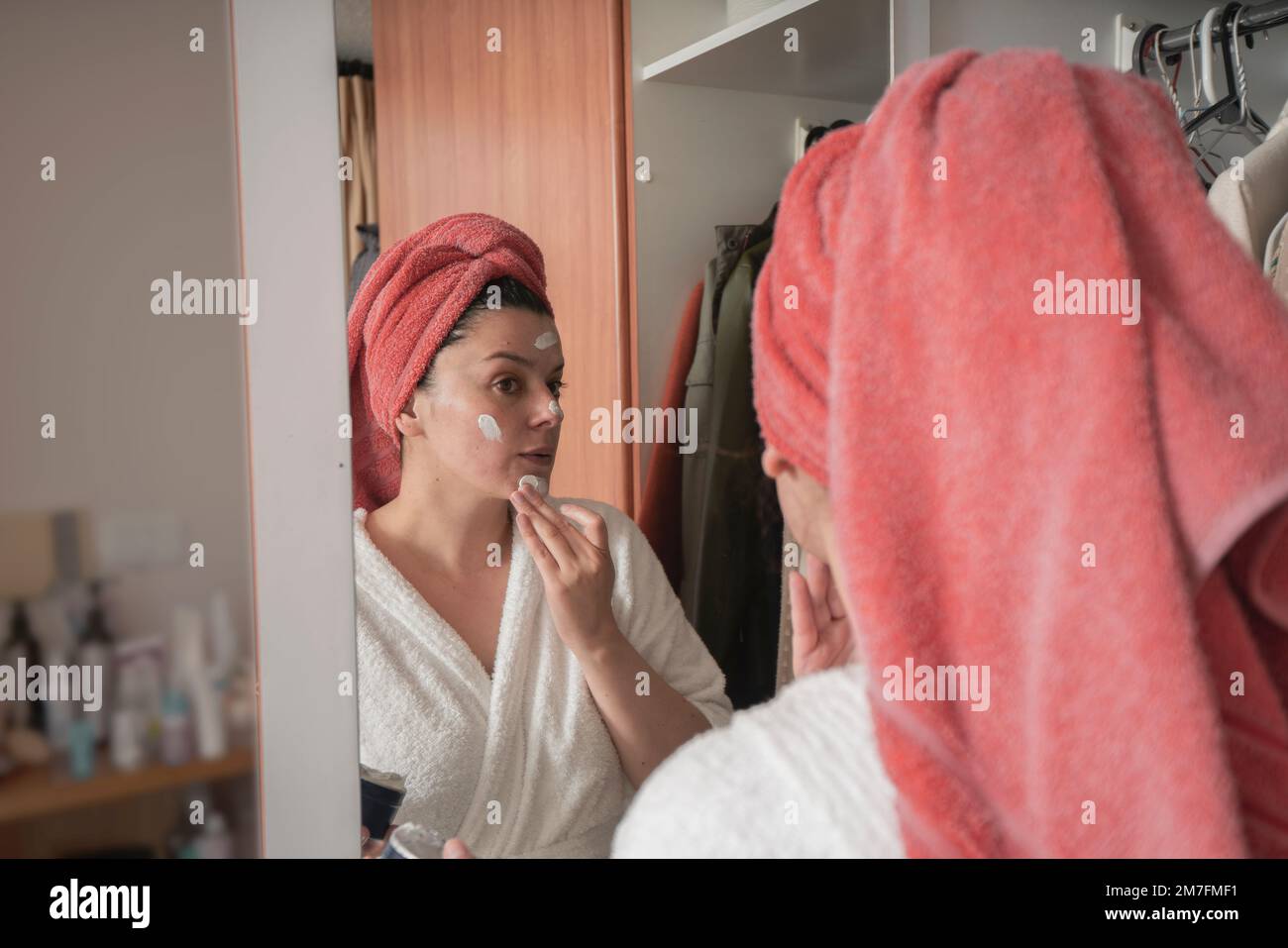 Belle femme hispanique dans un peignoir blanc et serviette rouge dans ses cheveux appliquant le maquillage avec sa main sur son visage devant le miroir sur le placard Banque D'Images