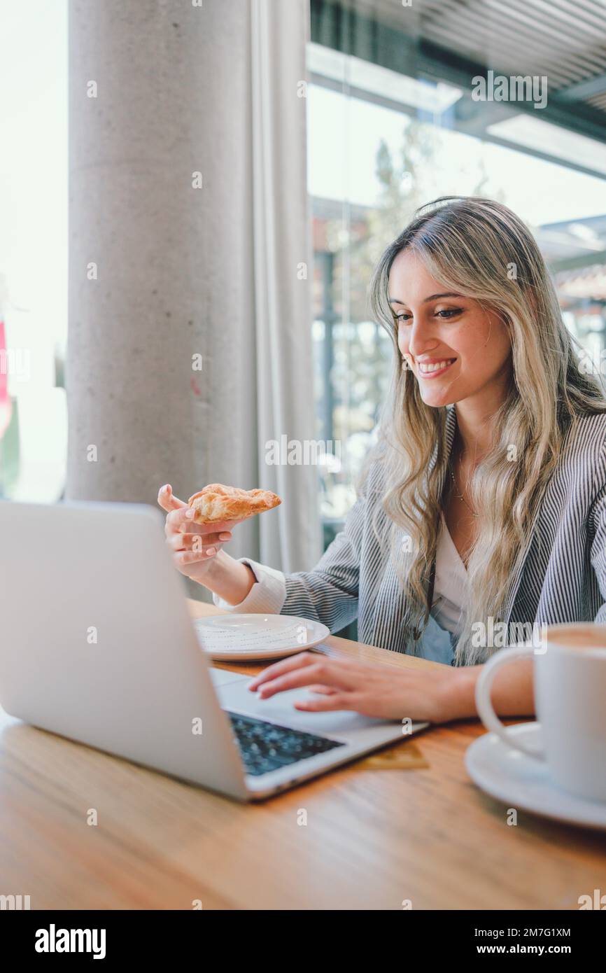 Jeune femme de direction du millénaire portant un blazer, mangeant un croissant et utilisant son ordinateur à l'intérieur Banque D'Images