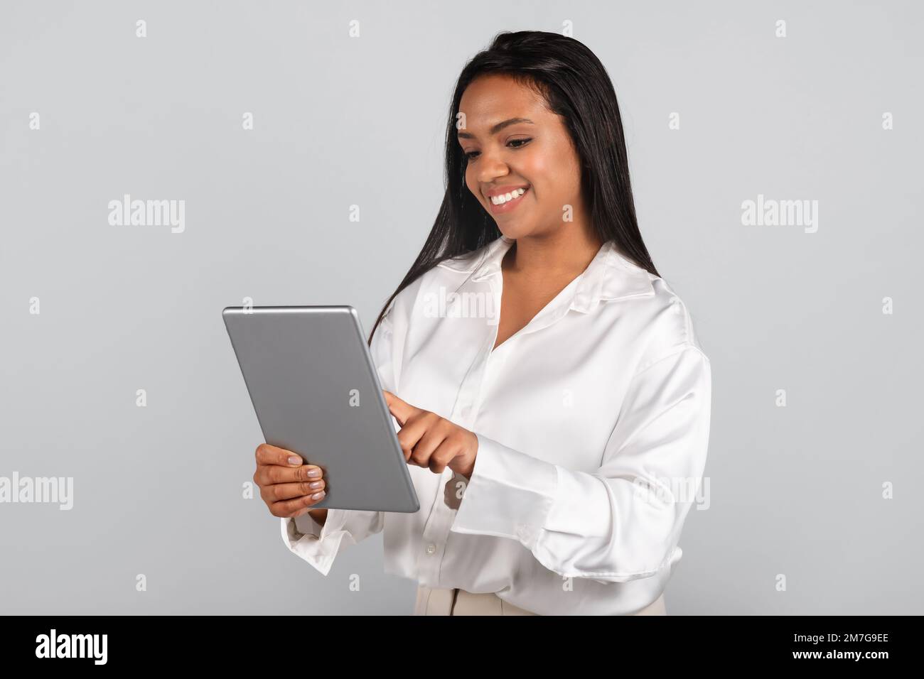 Gaie et confiante femme d'affaires noire millénaire en blouse blanche dactylographiant sur une tablette Banque D'Images