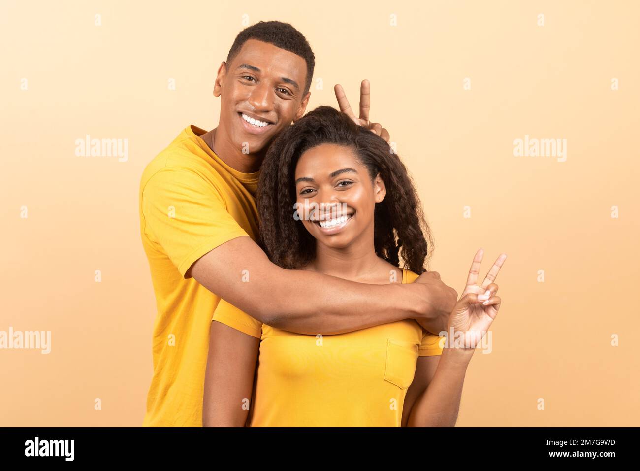 Joyeux jeune homme noir embrassant sa petite amie et faisant des cornes avec les doigts, posant ensemble sur fond jaune studio Banque D'Images
