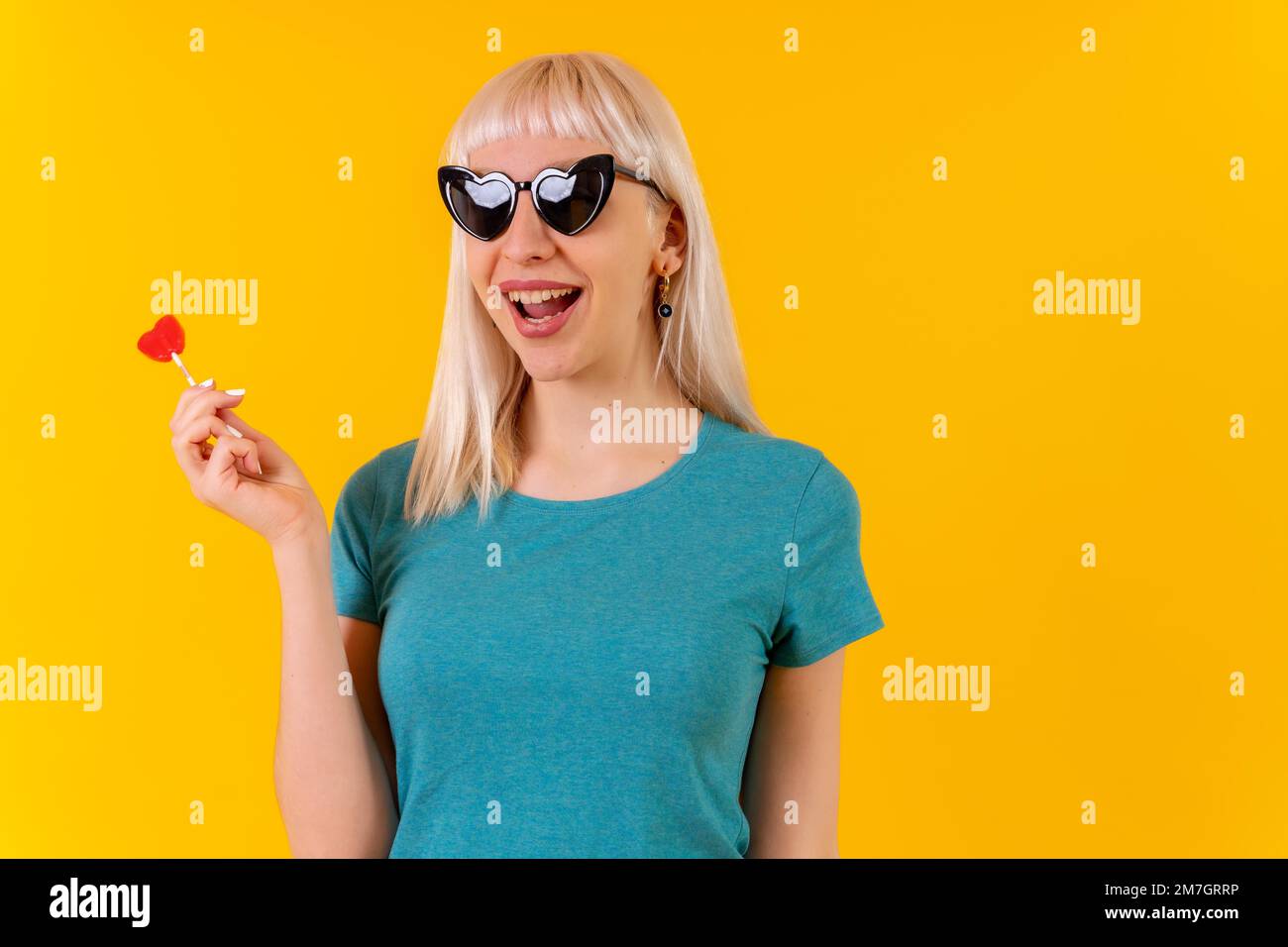 Manger un coeur Lollipop, blonde caucasienne fille en studio sur fond jaune Banque D'Images