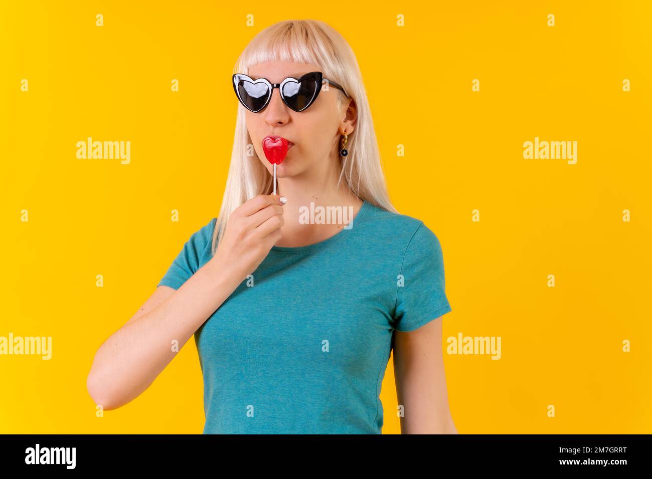 Manger un coeur Lollipop, blonde caucasienne fille en studio sur fond jaune Banque D'Images