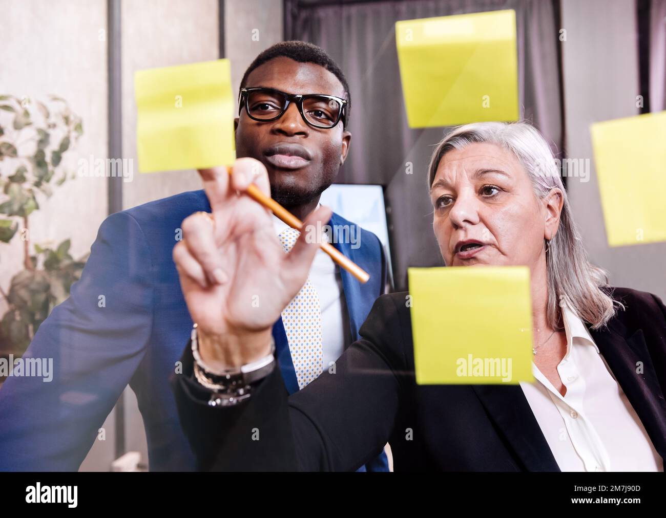 Femme d'affaires mûre pointant vers des notes adhésives et expliquant des données à des collègues noirs tout en se tenant derrière une fenêtre dans un bureau moderne Banque D'Images