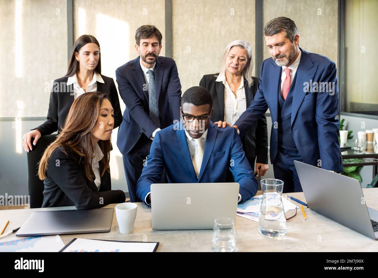 Homme d'affaires noir sérieux en costume et lunettes de vue parcourant les données sur netbook tout en étant assis à table parmi des collègues multiraciaux au bureau Banque D'Images