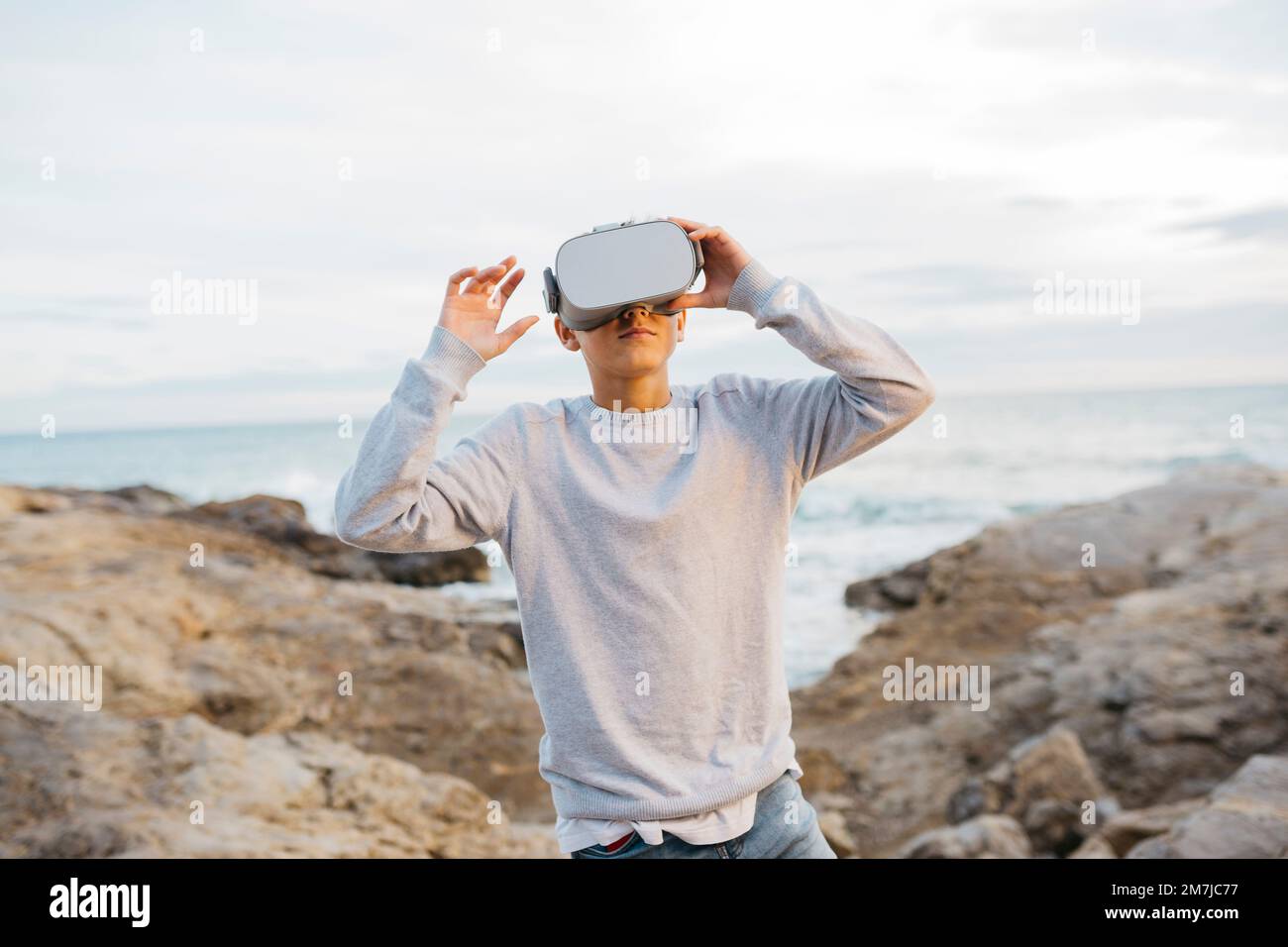 Garçon portant un simulateur de casque de réalité virtuelle sur un brise-lames de roche, à côté de la mer Banque D'Images