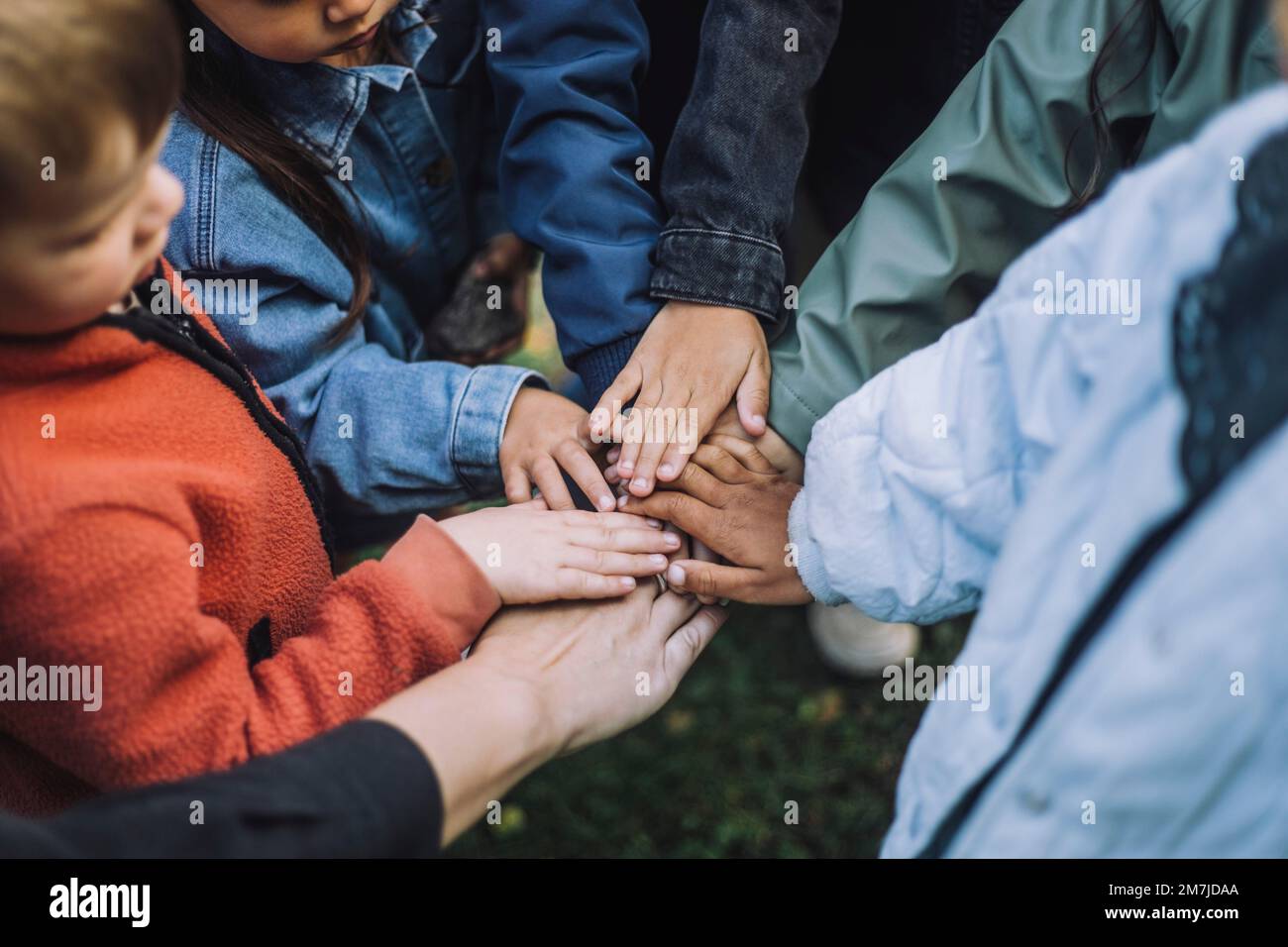 Des élèves préscolaires multiraciaux joueurs empilent les mains avec un enseignant dans le parc Banque D'Images