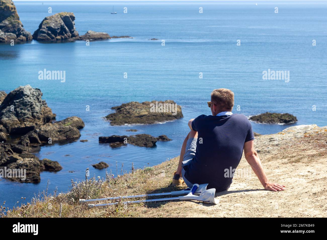 Homme handicapé méconnaissable assis sur le rocher avec ses béquilles à côté, tout en se reposant et en appréciant le magnifique paysage marin breton Banque D'Images