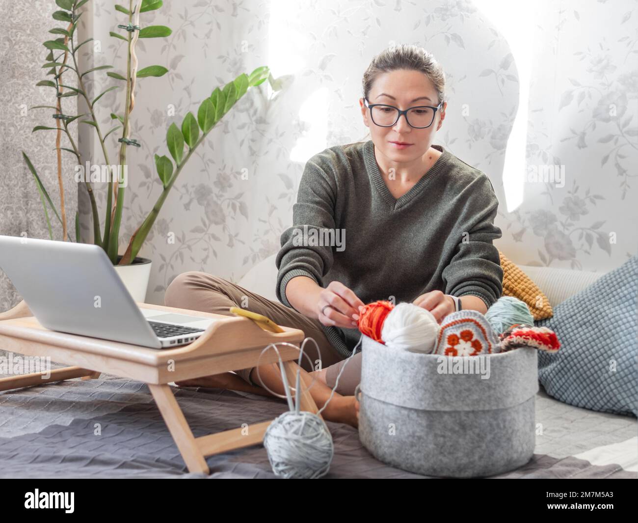 Femme choisit le fil pour crotcheting avec le film sur ordinateur portable. Apprenez à coudre à partir de leçons vidéo sur Internet. Maison confortable éclairée par la lumière du soleil. Banque D'Images