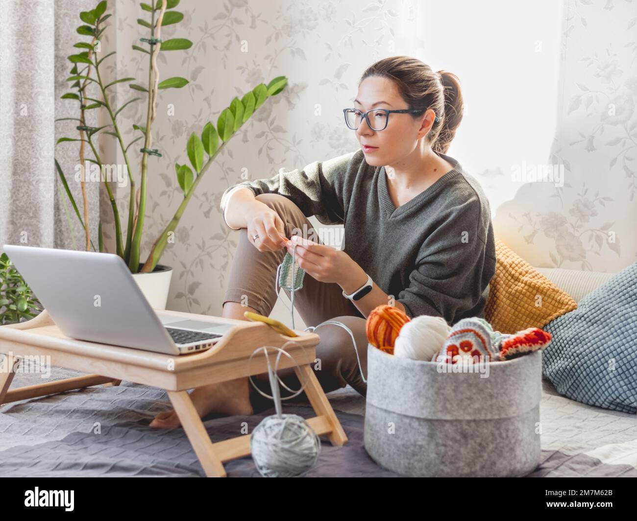 Une femme fait du crocheting tout en regardant un film sur un ordinateur portable. Apprenez à coudre à partir de leçons vidéo sur Internet. Maison confortable éclairée par la lumière du soleil. Banque D'Images