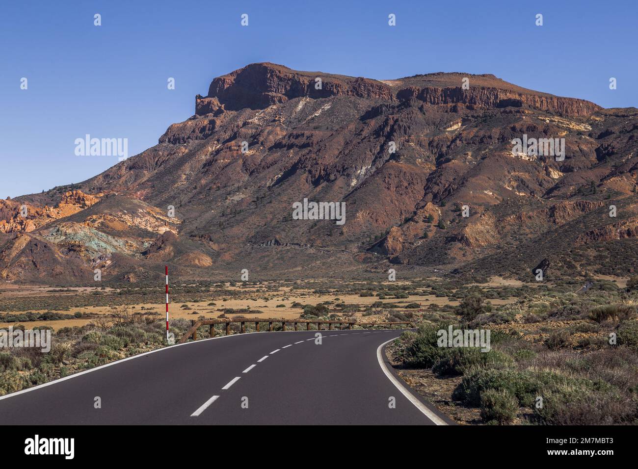 Une courbe sur l'autoroute en vue d'une énorme roche brune, noire et orange, avec la base jaune recouverte de buissons et d'arbustes verts, en dessous Banque D'Images