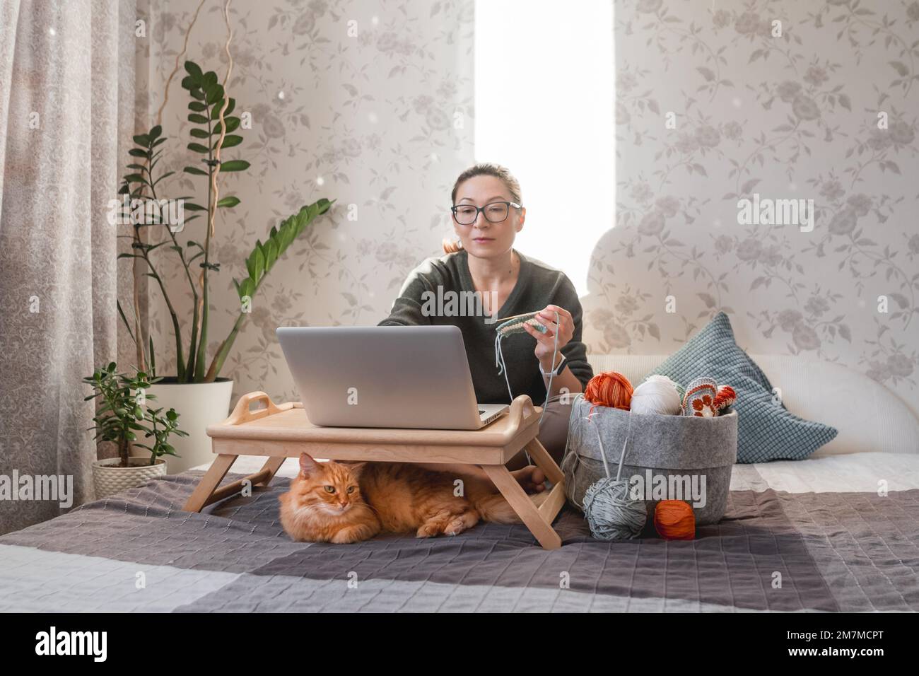 Une femme fait du crocheting tout en regardant un film sur un ordinateur portable. Apprenez à coudre à partir de leçons vidéo sur Internet. Maison confortable éclairée par la lumière du soleil. Banque D'Images