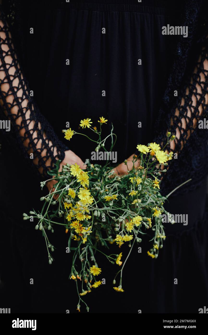 Femme vêtue de noir tenant des fleurs sauvages jaunes derrière son dos Banque D'Images