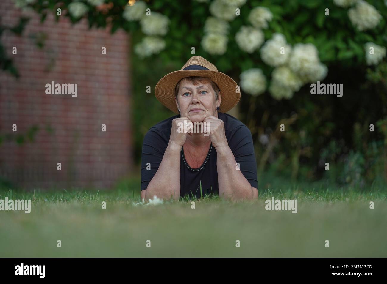 Une jeune grand-mère dans un chapeau de paille repose sur l'herbe dans le jardin avec son menton sur ses mains, vue du sol Banque D'Images