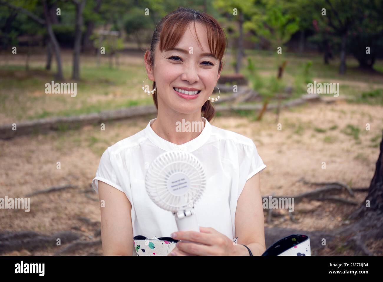 Une femme japonaise mûre à l'extérieur dans un parc lors d'une journée chaude tenant un petit ventilateur électrique. Banque D'Images