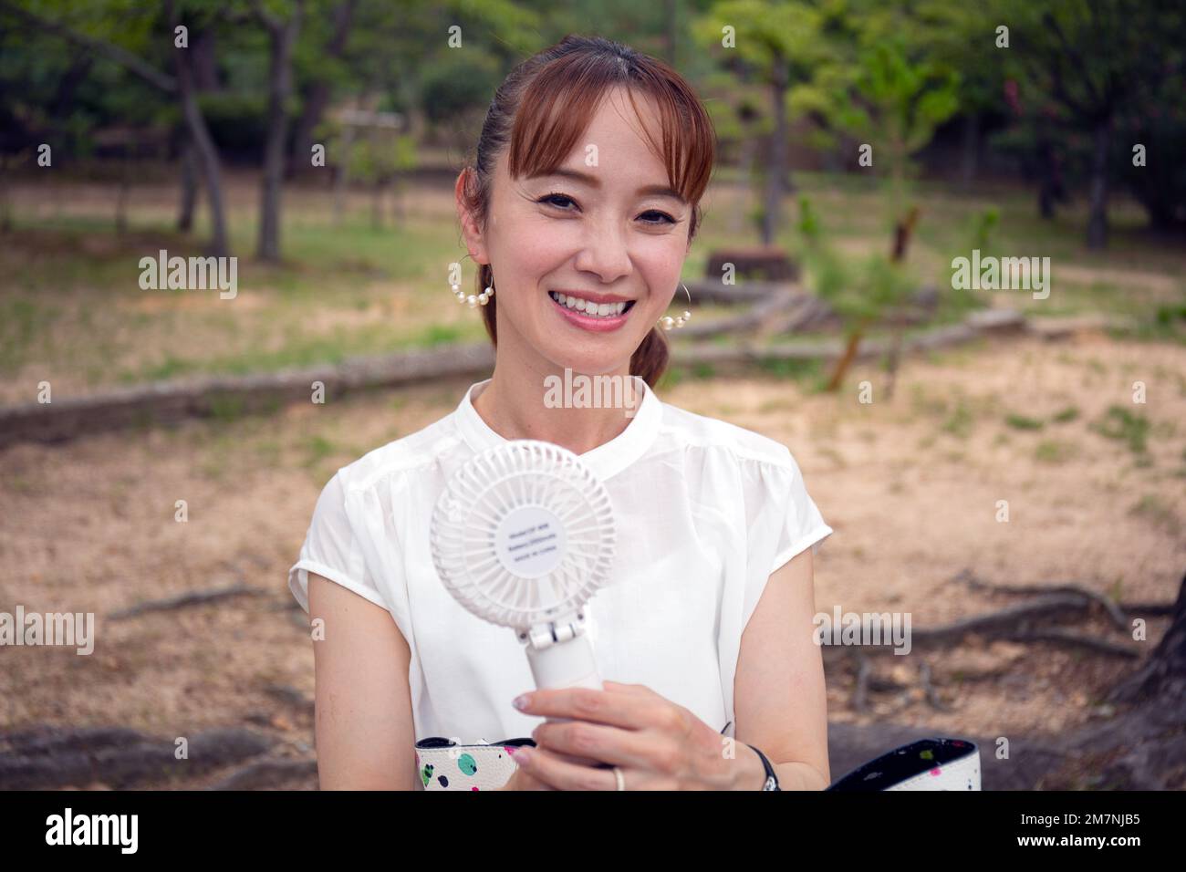 Une femme japonaise mûre à l'extérieur dans un parc lors d'une journée chaude tenant un petit ventilateur électrique. Banque D'Images