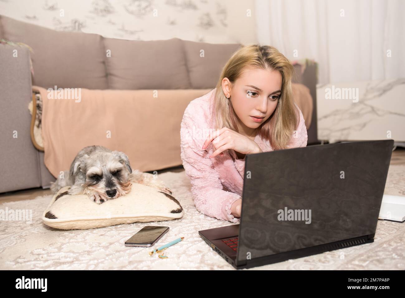 Une jeune femme blonde travaille sur un ordinateur portable, un chien gris est allongé sur la moquette à côté d'elle. Banque D'Images