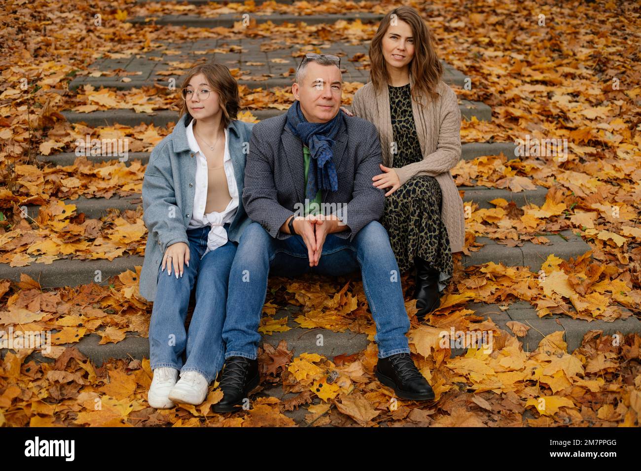 Une famille heureuse de trois personnes s'assoit dans un escalier recouvert de feuilles jaunes tombées. Les parents et les adolescents marchent dans le parc d'automne. Banque D'Images