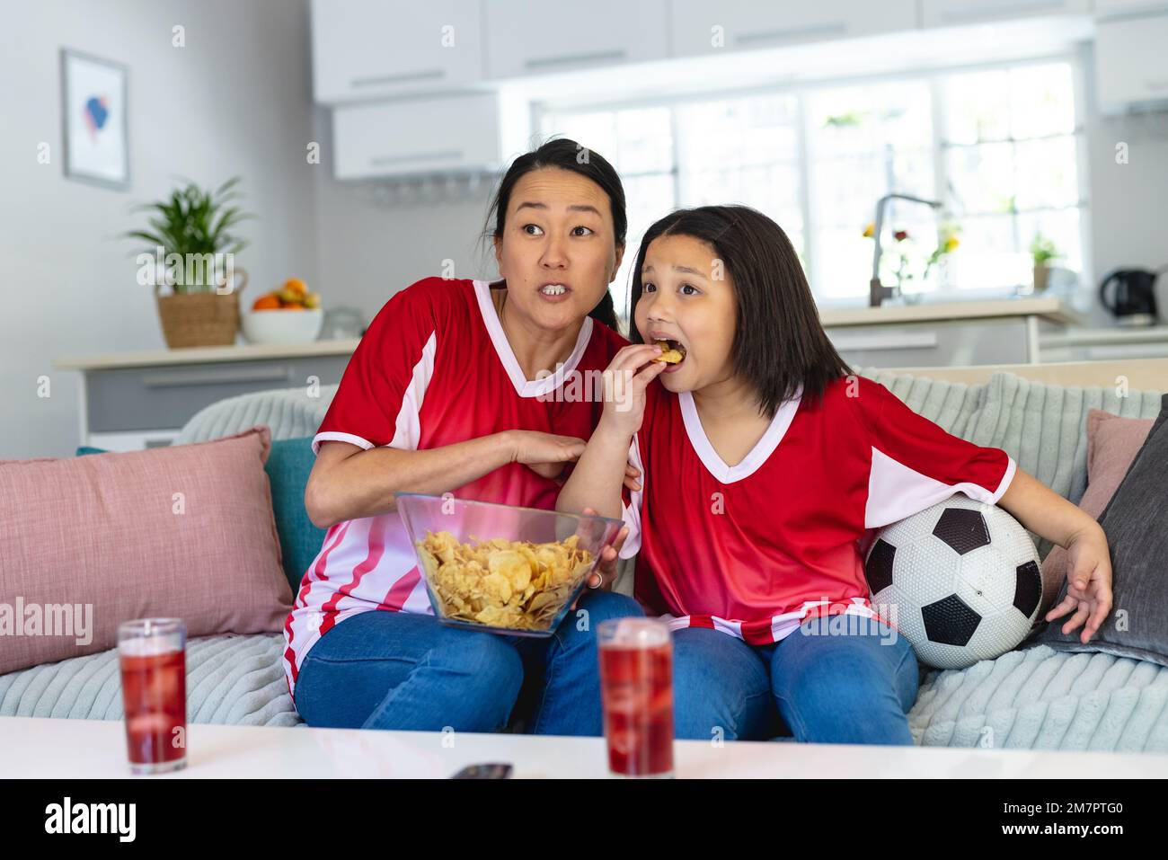 La mère et la fille asiatiques dans des maillots de football regardant le match de football, soutenant et mangeant des chips Banque D'Images