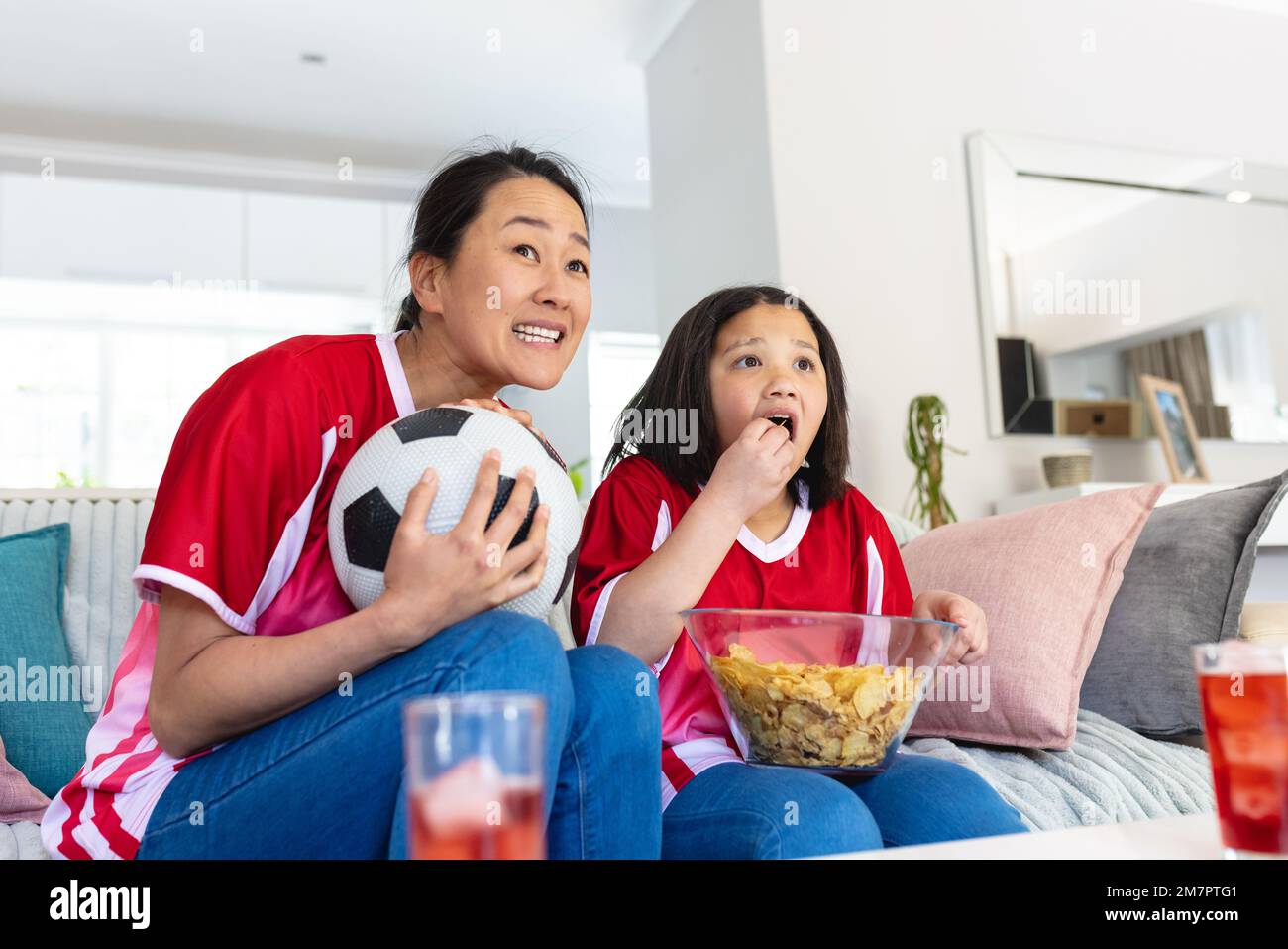 La mère et la fille asiatiques dans des maillots de football regardant le match de football, soutenant et mangeant des chips Banque D'Images