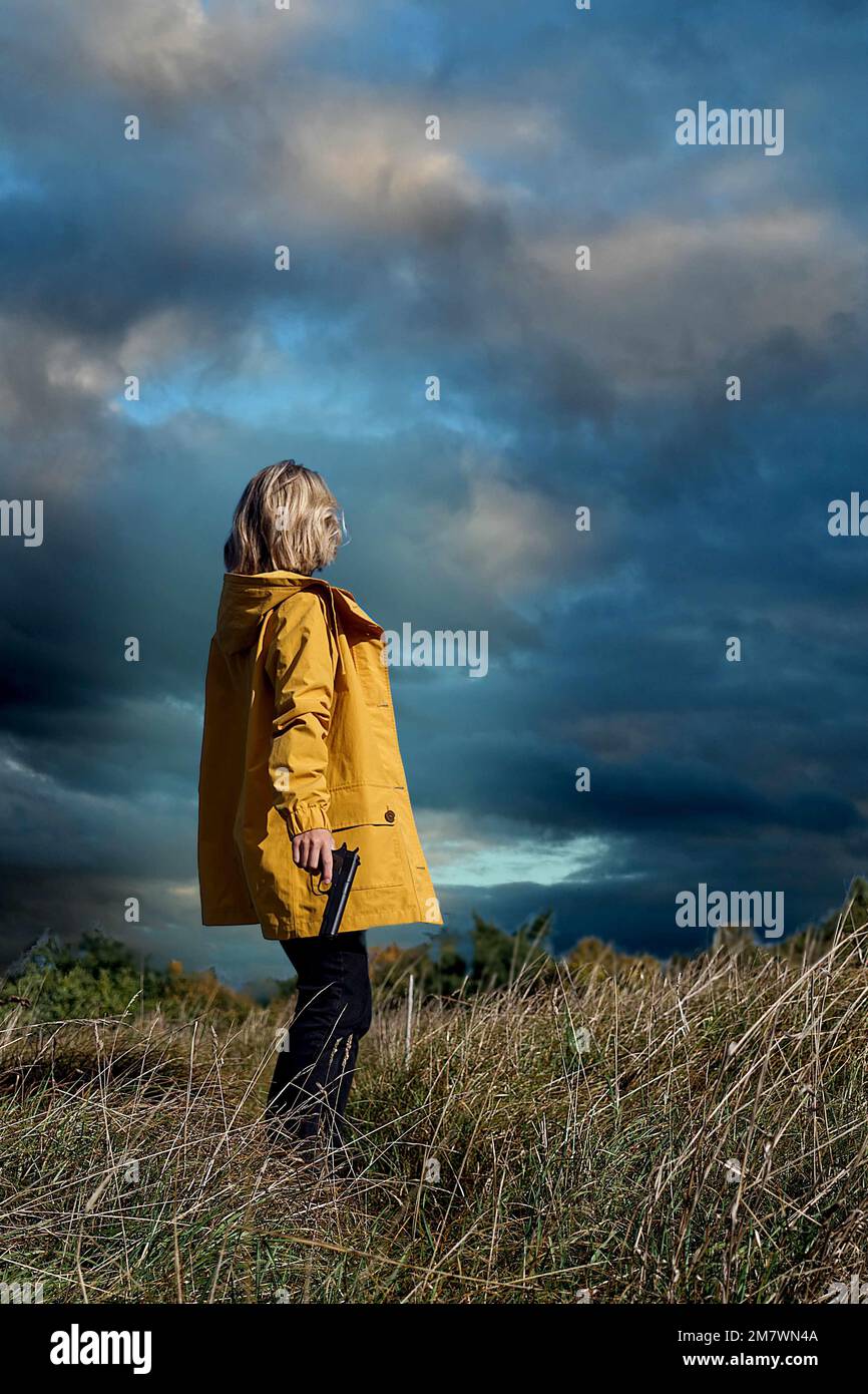 femme blonde aux cheveux mi-longueur et portant une veste jaune portant une arme à feu dans le pays. style de couverture de livre de thriller. Banque D'Images