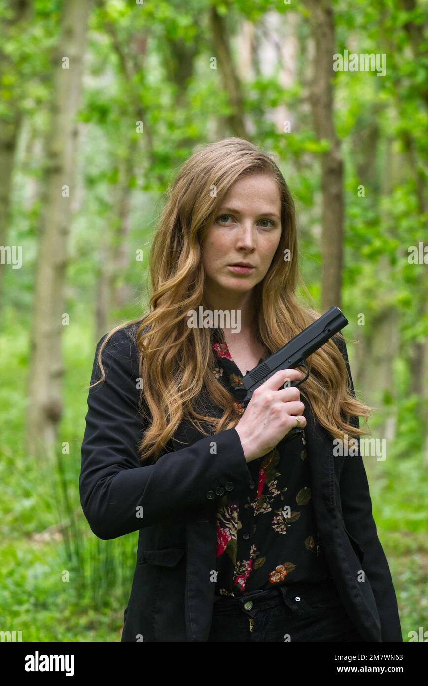Jeune femme aux cheveux rouges vêtue de noir et d'un pistolet dans les bois, thriller, mystère, style couverture de livre d'aventure. Banque D'Images