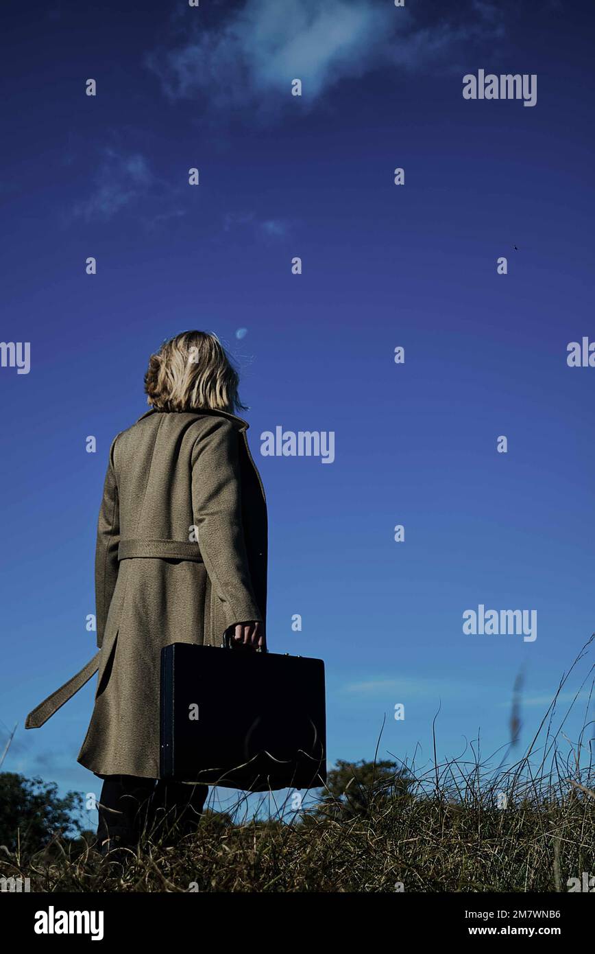 Femme blonde en manteau vert long portant un Briefcase dans la prairie. Style de couverture de livre de thriller d'espion mystère Banque D'Images