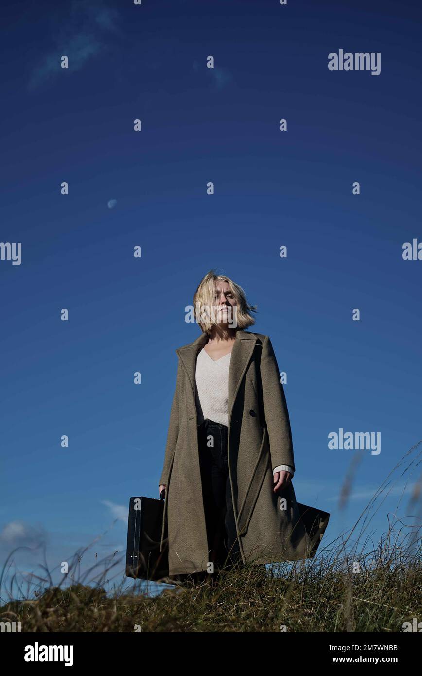 Femme blonde en manteau vert long portant un Briefcase dans la prairie. Style de couverture de livre de thriller d'espion mystère Banque D'Images