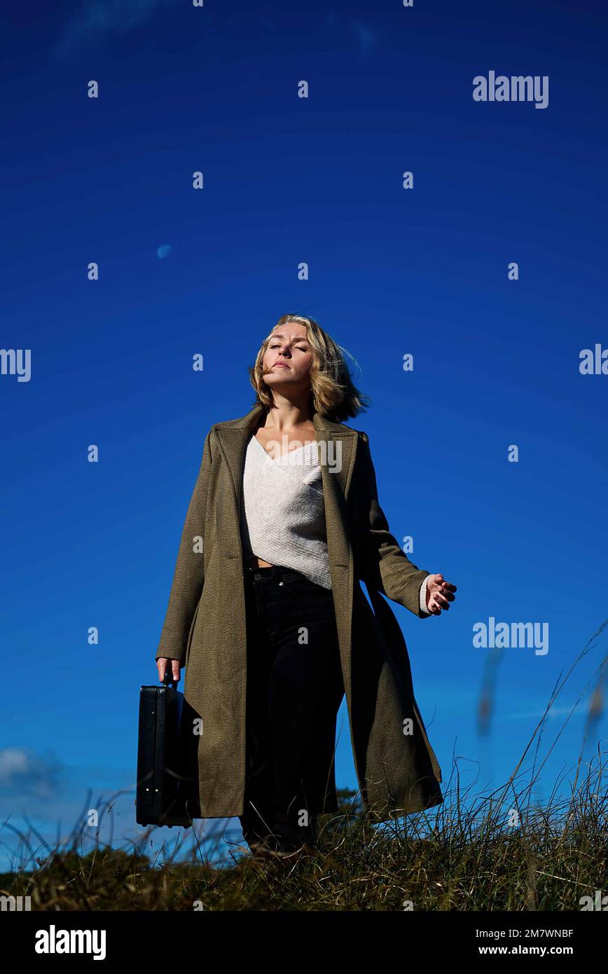 Femme blonde en manteau vert long portant un Briefcase dans la prairie. Style de couverture de livre de thriller d'espion mystère Banque D'Images