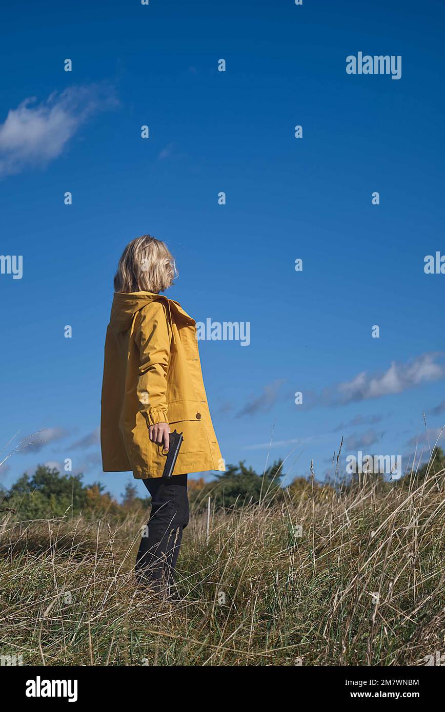 femme blonde aux cheveux mi-longueur et portant une veste jaune portant une arme à feu dans le pays. style de couverture de livre de thriller. Banque D'Images