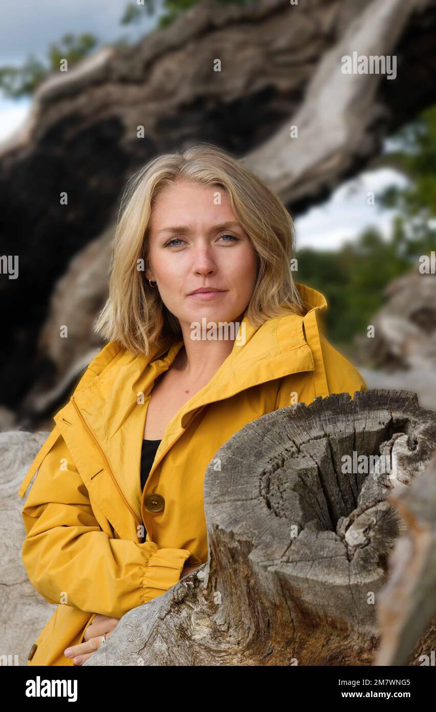 Une jeune femme blonde sous un manteau jaune debout contre un chêne mort tombé. Banque D'Images