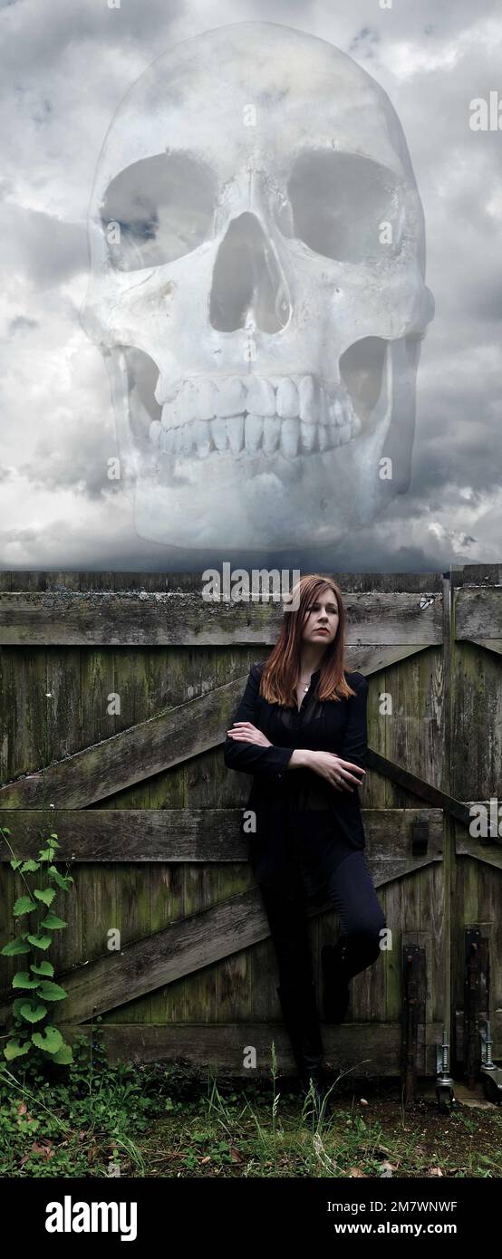 Jeune femme en noir se penchait contre une vieille porte avec un crâne fantomatique dans les nuages au-dessus d'elle. Banque D'Images