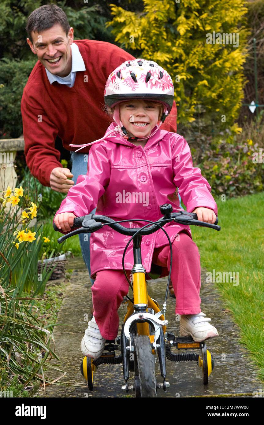 Un homme qui aide une jeune fille à faire son vélo jaune avec des stabilisateurs sur la voie dans un jardin Banque D'Images