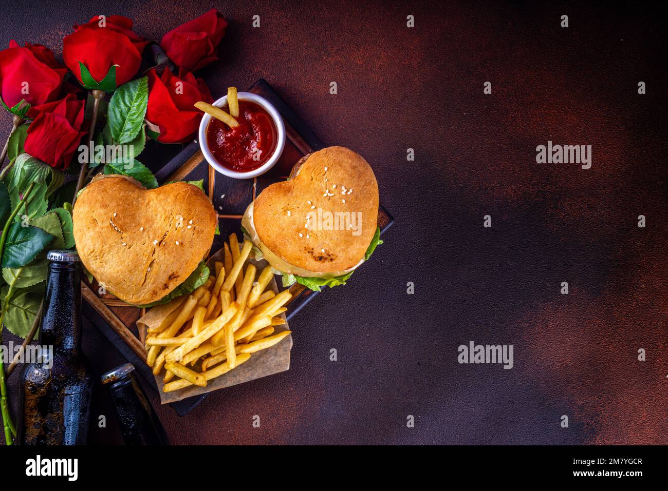 Hamburgers en forme de cœur pour la Saint-Valentin. Deux savoureux cheeseburgers avec frites et bouteilles de bière sur fond de table sombre. Idée pour le dîner de la Saint-Valentin Banque D'Images