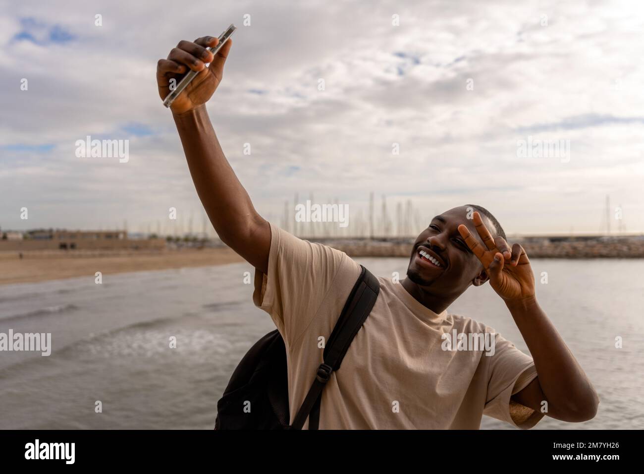 Joyeux jeune Africain américain dans des vêtements décontractés prenant selfie sur smartphone tout en montrant le signe de victoire près de la mer à la plage Banque D'Images