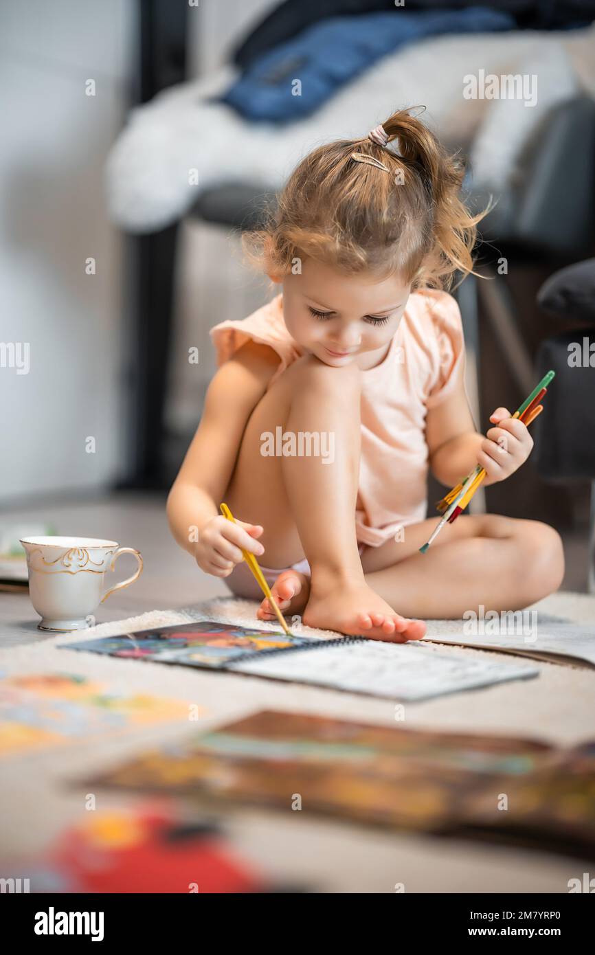 Petite fille s'assoit sur le sol à la maison et dessine avec des peintures et des pinceaux dans un livre de coloriage. Créativité et éducation de la petite enfance Banque D'Images