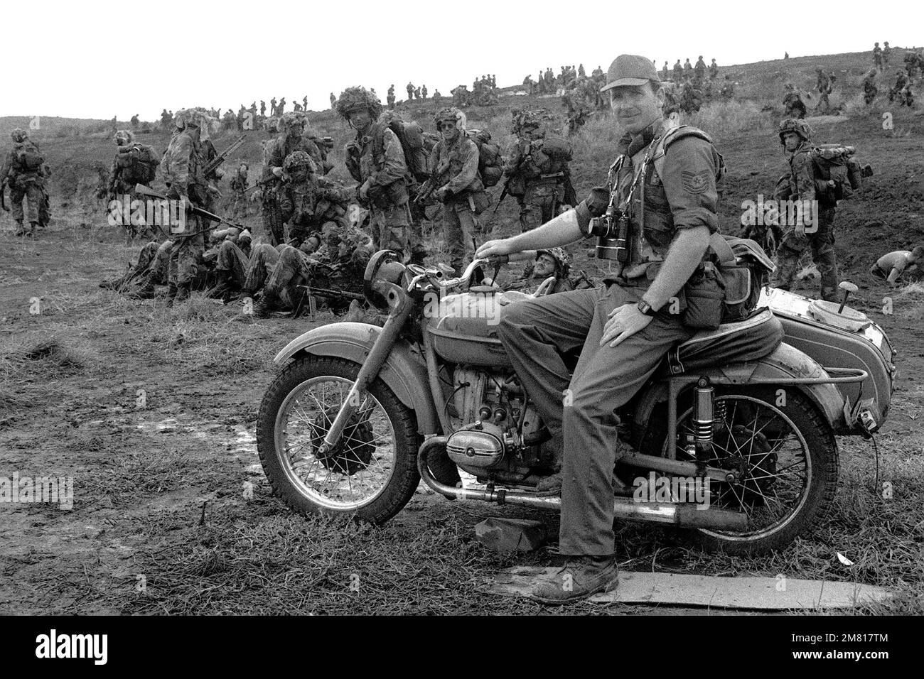 Un photographe de l'Armée de l'Air pose sur une moto à l'aérodrome de point Salines au cours de l'opération multinationale multiservice URGENT FURY. Se détendre sur la crête derrière lui sont membres de la 82nd Airborne Division. Objet opération/série: FUREUR D'URGENCE pays: Grenade (GRD) Banque D'Images