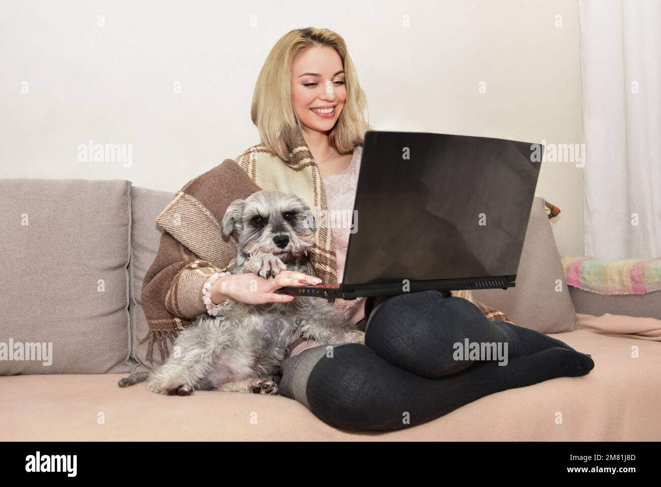 Jolie femme blonde sourit et embrasse un chien mignon sur le canapé tout en regardant des films sur un ordinateur portable. Banque D'Images