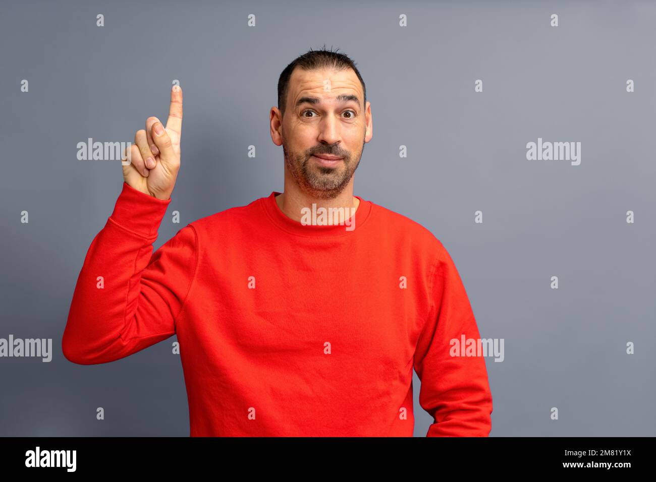 Portrait d'un homme hispanique barbu vêtu d'un pull-over rouge heureux excité pointant le doigt vers l'espace publicitaire isolé sur fond gris. Banque D'Images