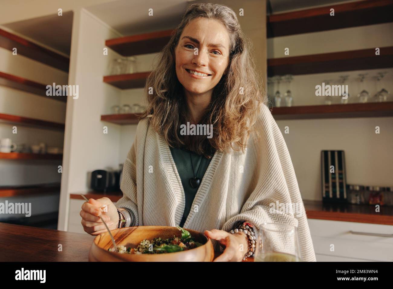 Femme mûre souriant à l'appareil photo tout en ayant un repas végétalien sain. Bonne femme âgée se servant elle-même un bol de bouddha sain à la maison. Femme prenant Banque D'Images