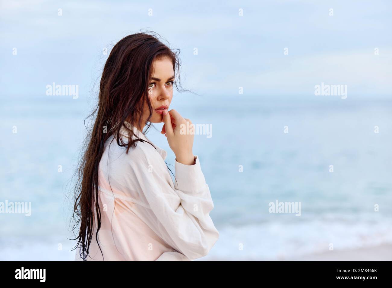 Portrait d'une belle femme pensive avec une peau bronzée dans une chemise de plage blanche avec des cheveux mouillés après avoir nagé sur la plage océan coucher de soleil lumière avec des nuages Banque D'Images