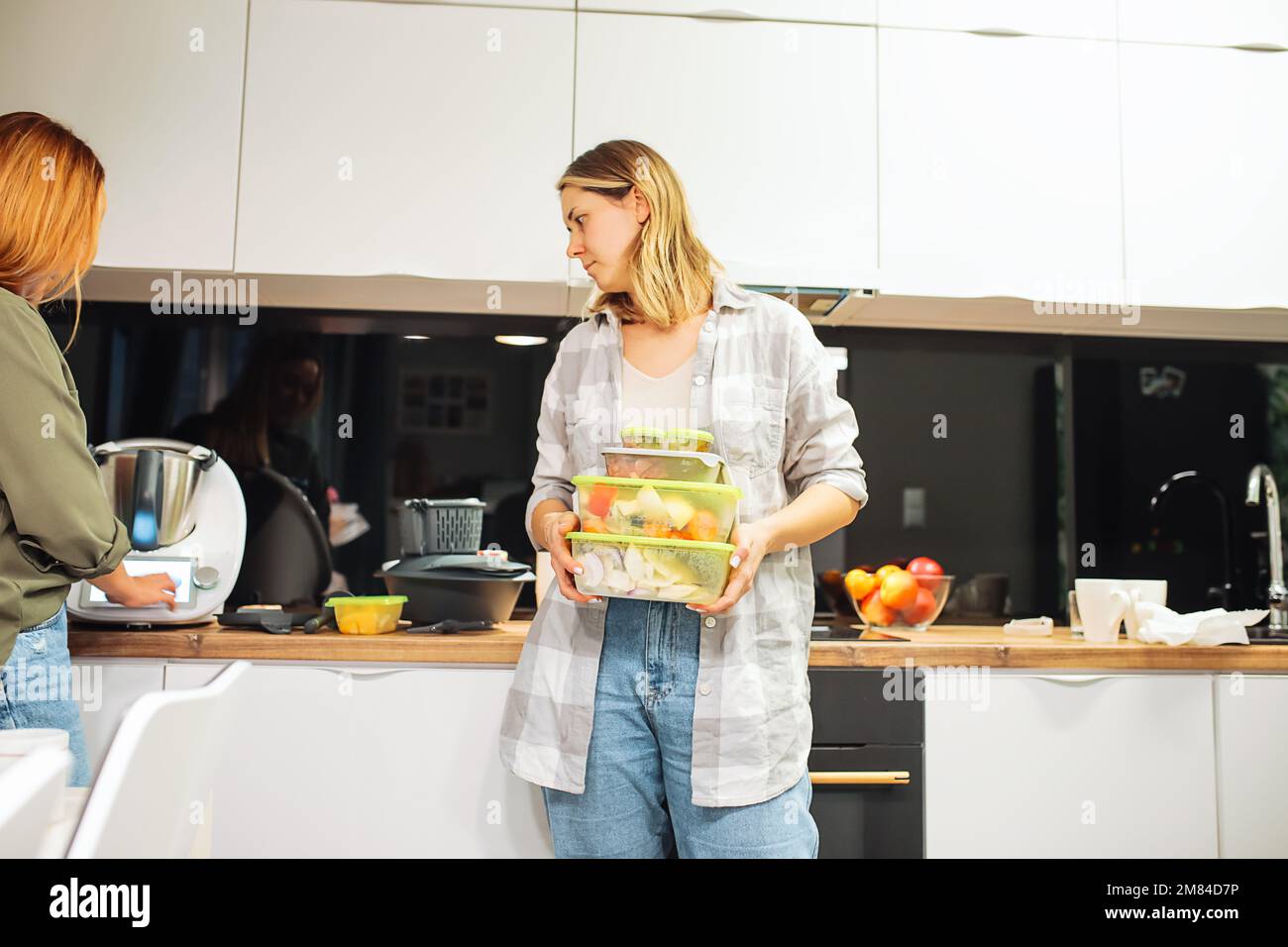 Une femme tient des boîtes en plastique avec des aliments et regarde les appareils électroniques modernes de cuisine. Les amis cuisinent ensemble à la maison. Banque D'Images