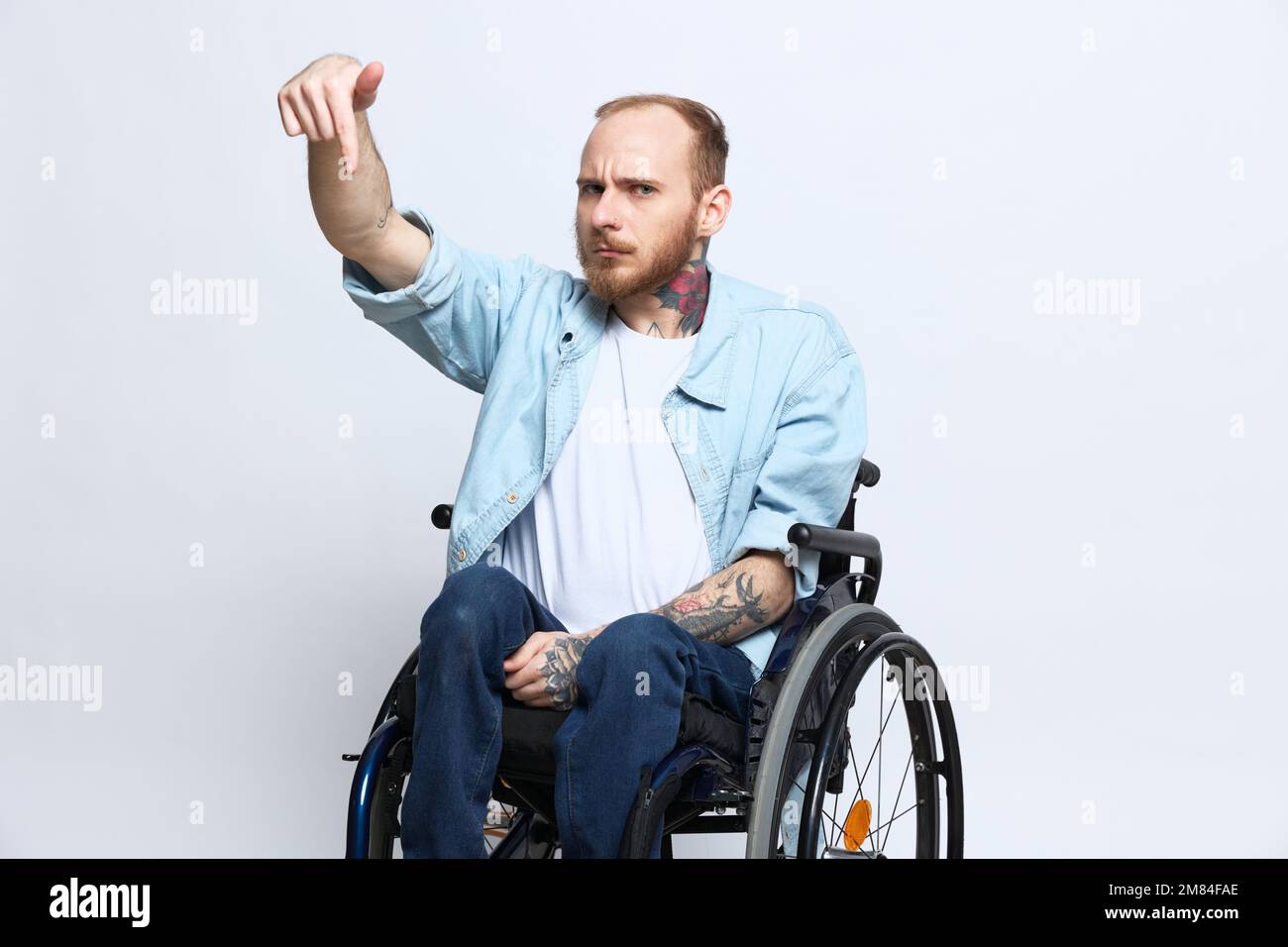Un homme dans un fauteuil roulant et pointant un doigt, un espace de copie, avec des tatouages sur ses mains est assis sur un fond gris studio, concept de santé une personne avec Banque D'Images