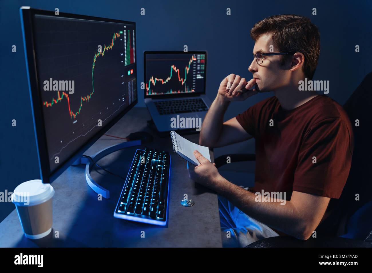 Crypto investisseur travaillant sur pc, notant les résultats de son analyse de marché de négociation, assis à table, regardant l'affichage avec concentration, l'analyse des mouvements de prix, l'ajustement de la stratégie d'investissement Banque D'Images
