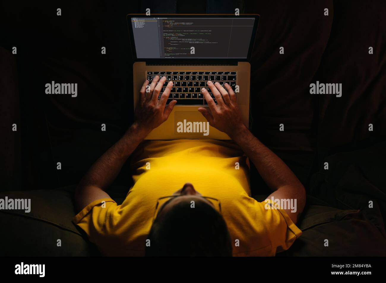 Un jeune programmeur tape du nouveau code sur un ordinateur portable tard dans la nuit, assis sur un canapé à la maison, travaillant des heures supplémentaires sur une nouvelle application. Concept de surtravail et de travail à distance. Vue de dessus Banque D'Images