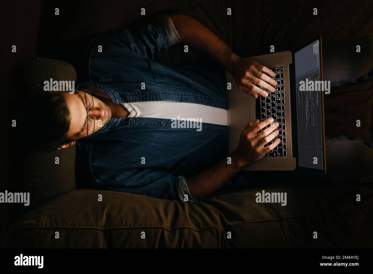 Vue de dessus d'un programmeur mâle posé sur un canapé et travaillant sur son ordinateur portable. Un codeur surtravaillé travaillant de chez lui la nuit. Banque D'Images