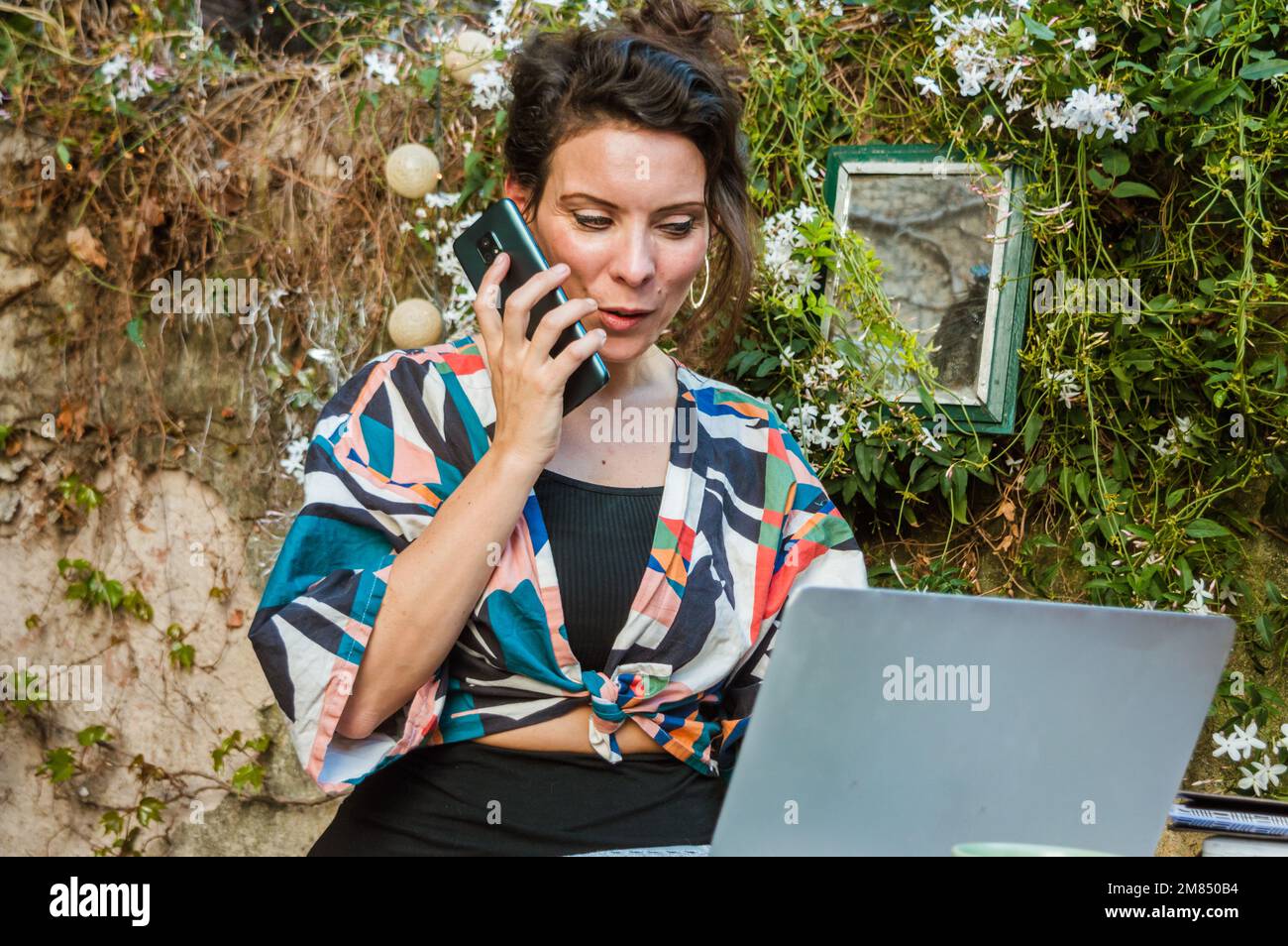 Bonne femme française caucasienne adulte travaillant avec son ordinateur portable sur la terrasse de sa maison, envoyant une note vocale avec le téléphone, communiquant avec sa cl Banque D'Images